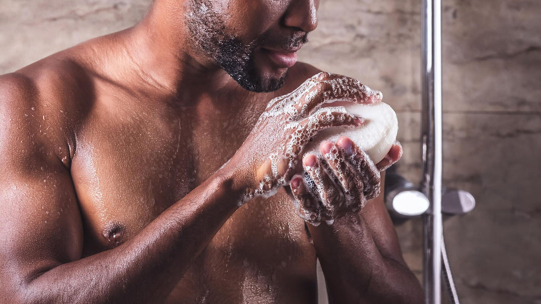 A shirtless man is taking a shower, holding a bar of soap lathered with foam in his hands. The background shows a tiled shower wall and a showerhead. The man appears focused on the soap as he washes himself.