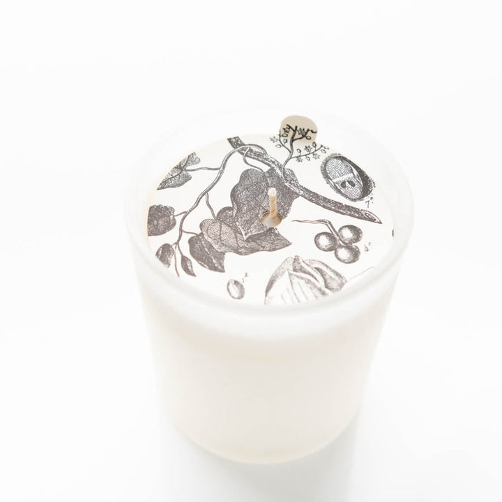 A white candle in a frosted glass holder. The candle surface features an intricate black and white illustration of leaves, fruits, and a tree branch surrounding the wick. The background is entirely white, highlighting the detailed artwork on the candle.