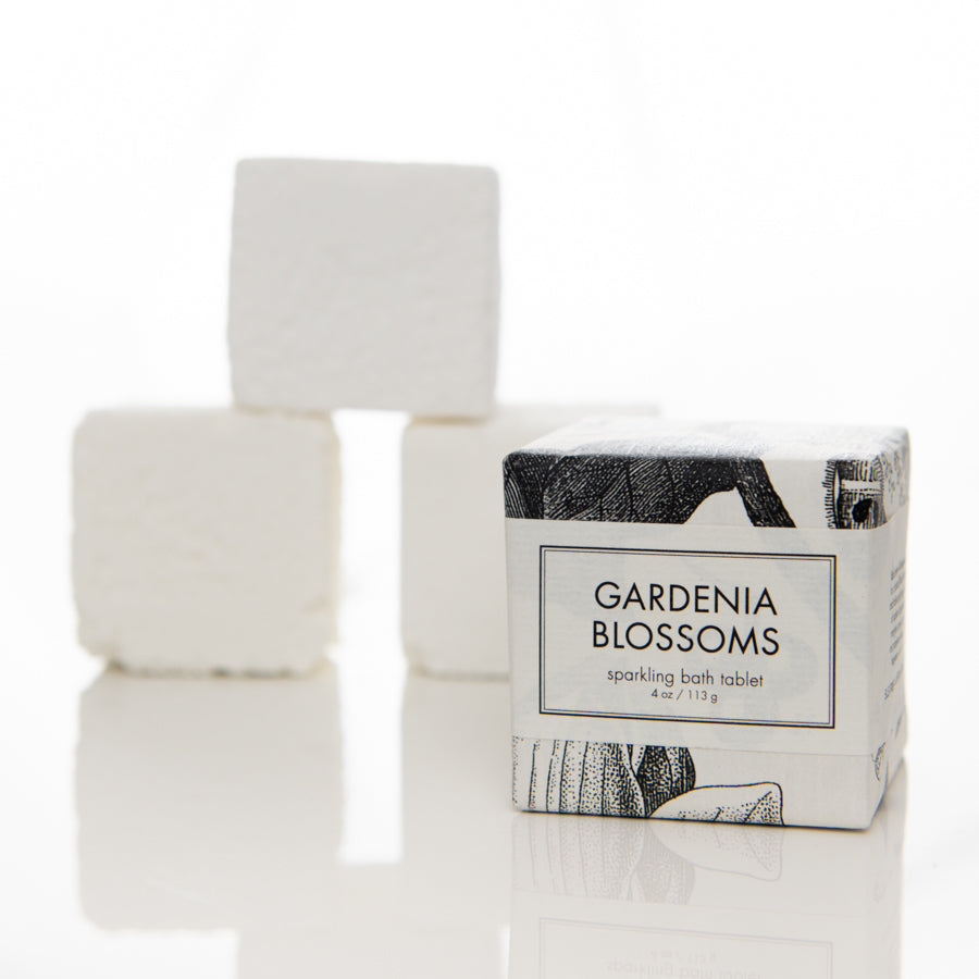 A sparkling bath tablet labeled "Gardenia Blossoms" stands in the foreground. Behind it, three unfocused white bath tablets are stacked on a reflective surface. The packaging features artistic, monochromatic floral designs.