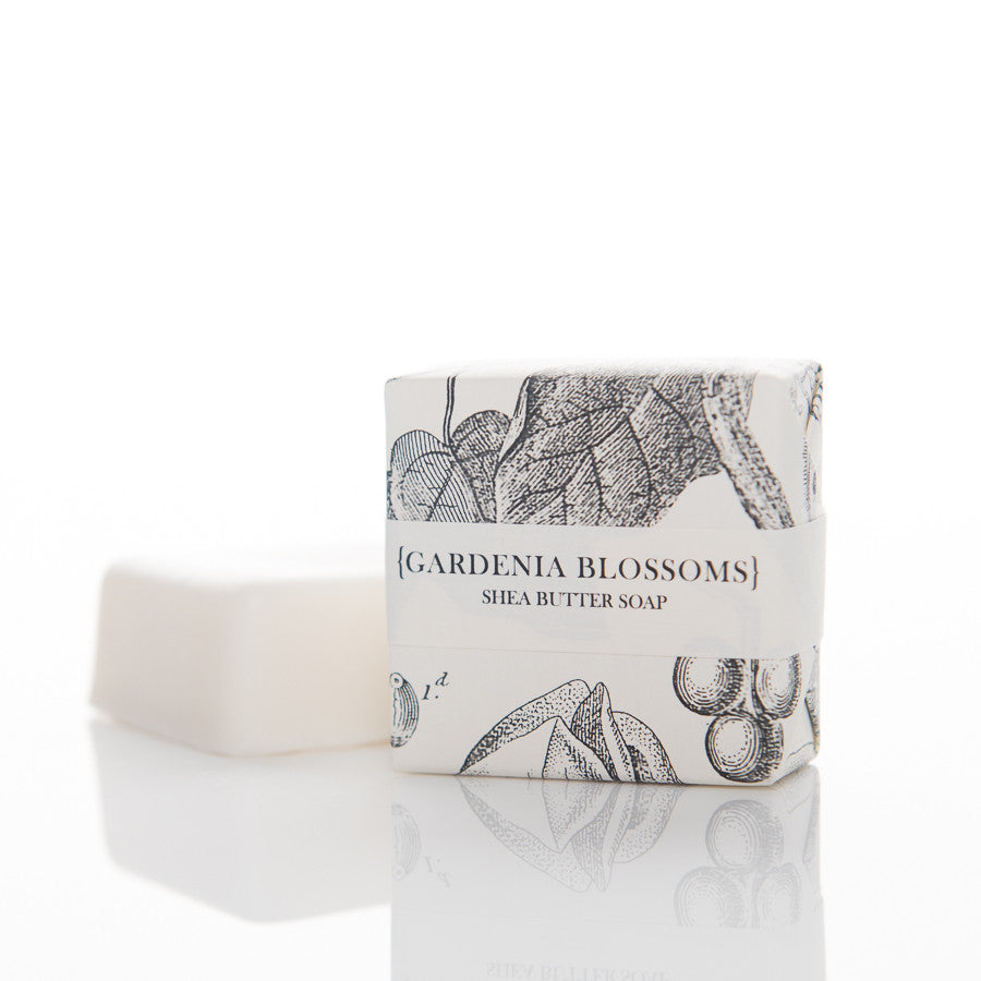 A bar of shea butter soap labeled "Gardenia Blossoms." The soap is partially unwrapped, revealing its white color. The wrapping paper features botanical illustrations of flowers and leaves in black and white. The scene is set against a white background.