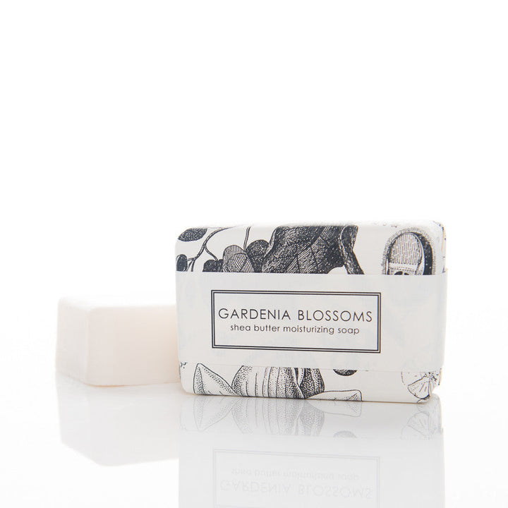 A rectangular bar of white soap is partially wrapped in elegant paper with floral designs. The label on the packaging reads "Gardnia Blossoms shea butter moisturizing soap." The soap bar is simple and unadorned, emphasizing a clean, minimalist look.