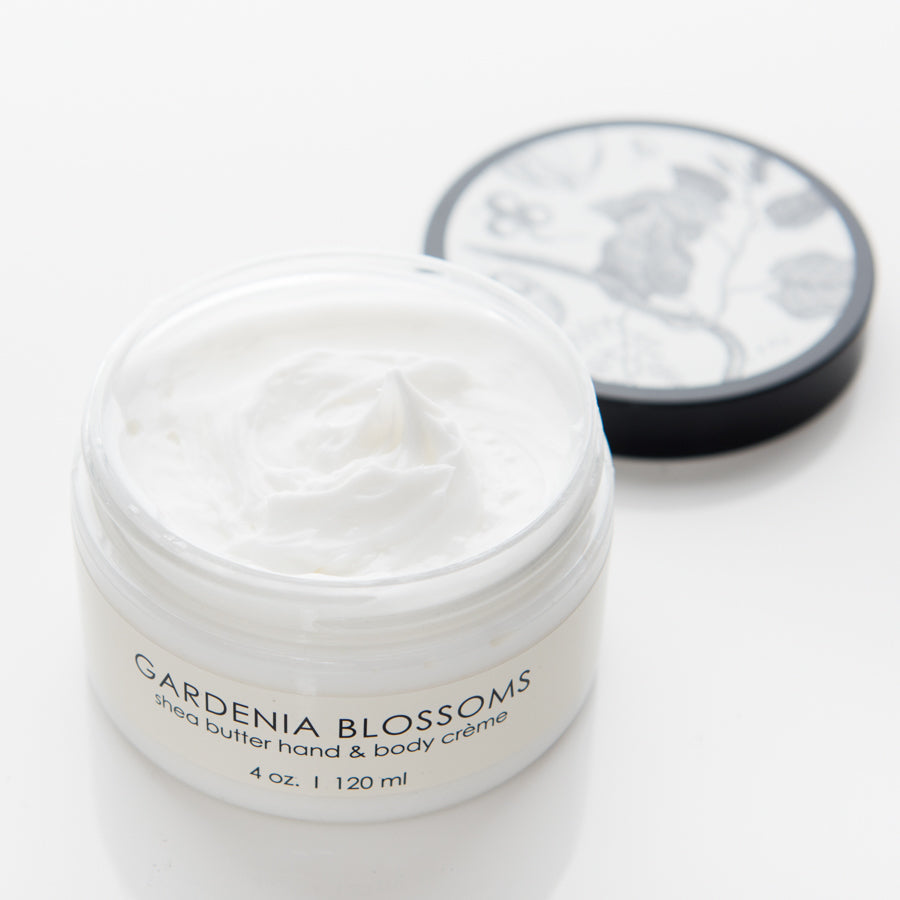 A white jar labeled "Gardenia Blossoms shea butter hand & body crème" is opened, showing a creamy white substance inside. The jar has a black lid with floral designs resting beside it. The container holds 4 oz. (120 ml) of the product.