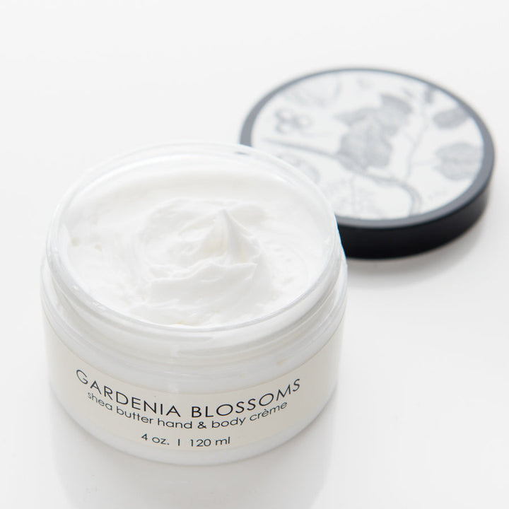 A white jar labeled "Gardenia Blossoms shea butter hand & body crème" is opened, showing a creamy white substance inside. The jar has a black lid with floral designs resting beside it. The container holds 4 oz. (120 ml) of the product.