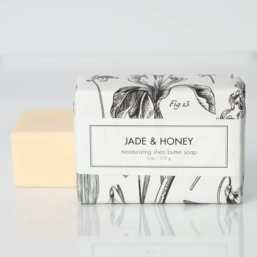 A wrapped bar of Formulary55's Jade and Honey Shea Butter Soap stands next to an unwrapped bar. The packaging features a black-and-white floral design with the soap's name and weight, 6 oz / 170 g, printed on the front. The unwrapped shea butter soap bar is beige and rectangular.