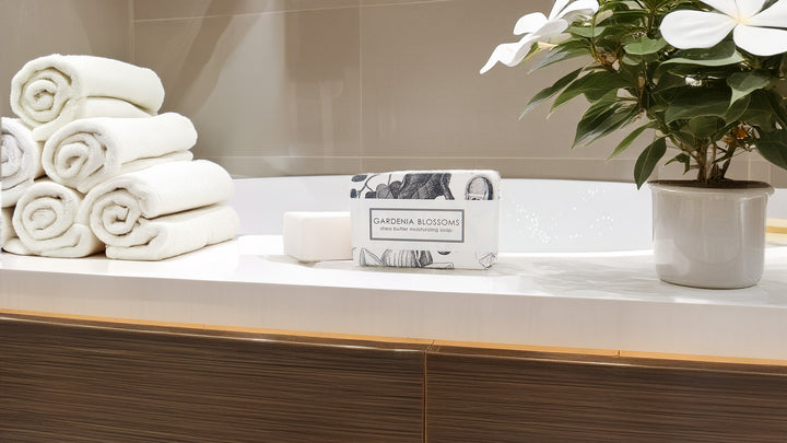 A modern bathroom features a stack of white rolled towels, a bar of soap, and a box labeled "Gardenia Blossoms" placed on a sleek countertop. A potted plant with white flowers adds a touch of greenery next to a luxurious white bathtub.