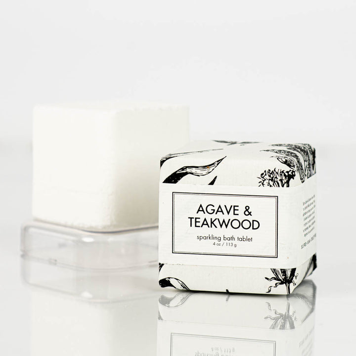 A white sparkling bath bomb tablet from Formulary55, partially wrapped in stylish paper adorned with black botanical illustrations, is pictured with the label "agave and teakwood." The tablet weighs 4 oz (113g) and is set against a plain background.