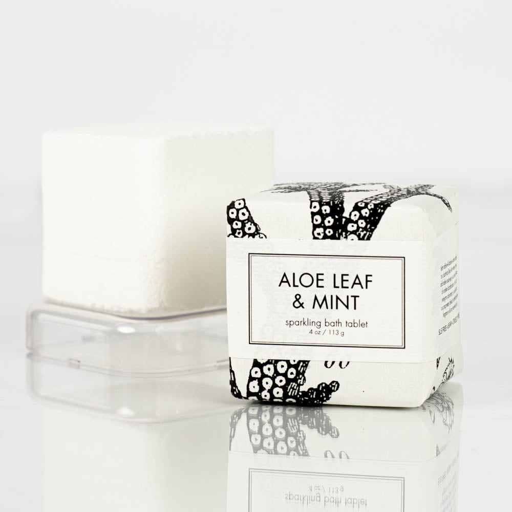 A Formulary55 Aloe Leaf and Mint Sparkling Bath Bomb Tablet, wrapped in white paper adorned with black designs, is placed beside an unwrapped white bath bomb on a clear plastic base. The packaging displays the text "Aloe Leaf &amp; Mint".