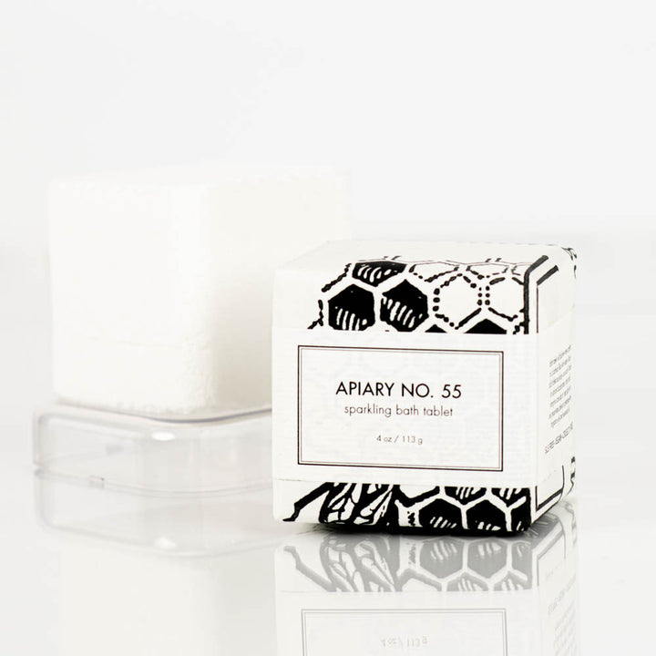 A handmade bath bomb labeled "Apiary Number 55 Sparkling Bath Bomb Tablet" by Formulary55 is displayed next to its packaging, which features a black and white design with honeycomb patterns and text that reads "APIARY NO. 55, sparkling bath tablet, 4 oz / 113 g".