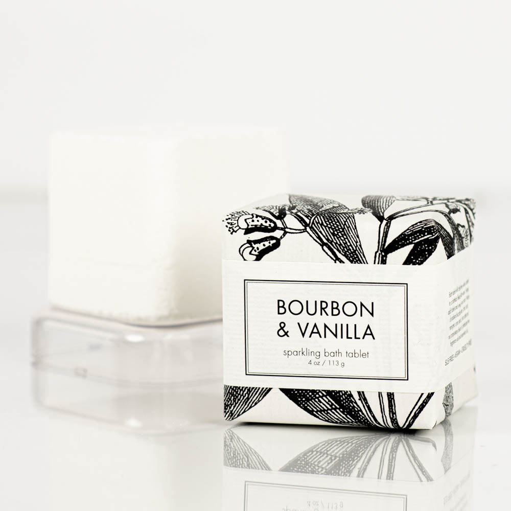 A Bourbon &amp; Vanilla Sparkling Bath Bomb Tablet from Formulary55, wrapped in packaging adorned with black and white botanical illustrations, is prominently displayed. Behind it, the unwrapped bath bomb—a simple white cube—sits elegantly on a clear stand. The overall look is both clean and sophisticated.