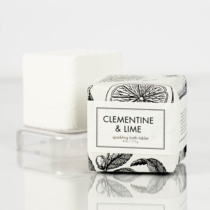 A square bath tablet labeled "clementine and lime Sparkling Bath Bomb Tablet by Formulary55, 4 oz / 113 g" is displayed with another tablet in the background. The packaging features black and white illustrations of citrus fruits and leaves. Both tablets rest on a clear surface.