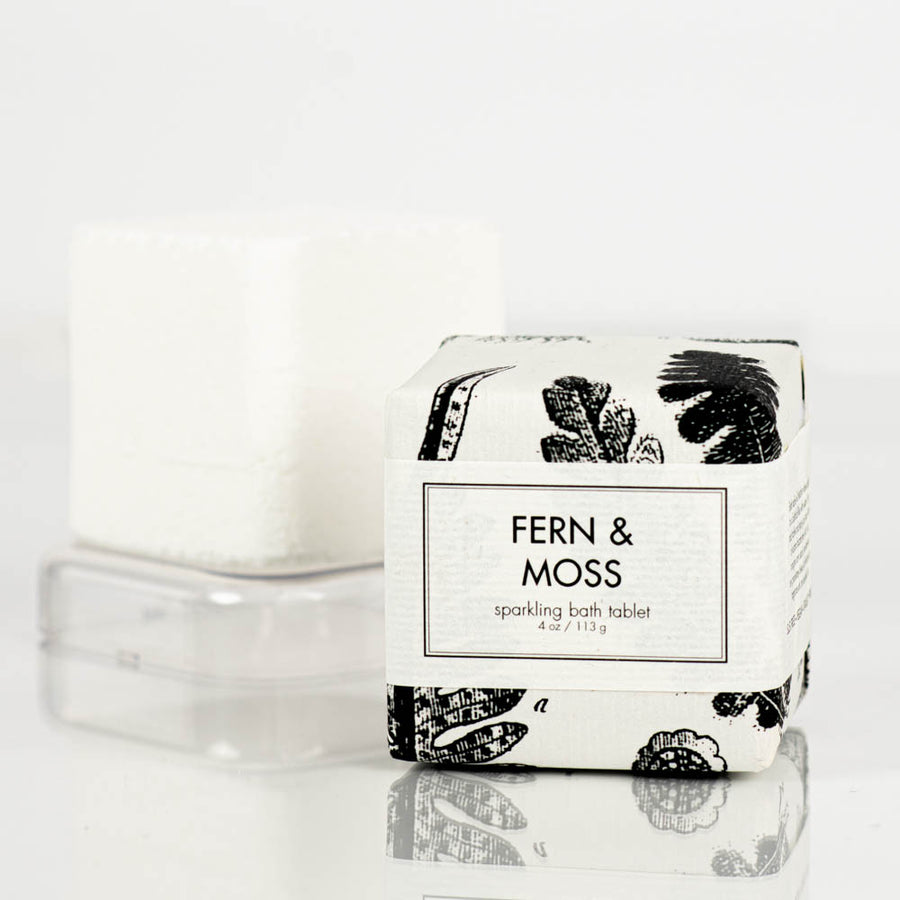 A white, sparkling bath bomb tablet wrapped in white paper adorned with black floral designs sits next to an unwrapped white bath bomb tablet on a small plastic tray. The packaging reads "Formulary55 fern and moss Sparkling Bath Bomb Tablet 4 oz / 113 g.