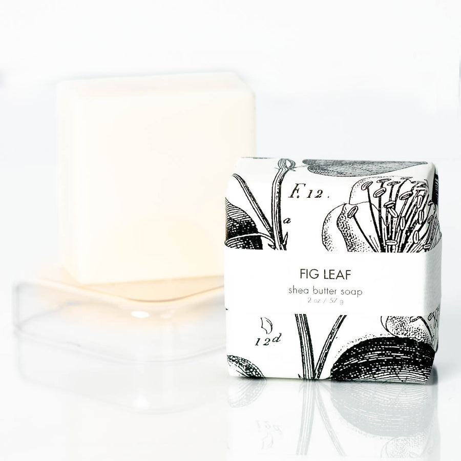 A bar of Formulary55's Fig Leaf natural petite shea butter soap is wrapped in white paper adorned with botanical sketches. The unwrapped natural soap bar sits on a clear tray behind the wrapped one, showcasing its creamy, off-white color.