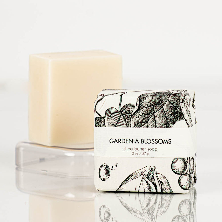 Close-up view of a natural shea butter bar soap in minimalist packaging. The label reads "gardenia blossoms petite shea butter natural bar soap" by Formulary55 and features a botanical illustration. The unwrapped soap bar is displayed on a clear soap dish in the background against a plain, white backdrop.