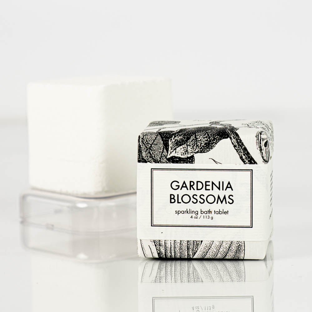 A square-shaped "gardenia blossoms Sparkling Bath Bomb Tablet" by Formulary55 is shown. The product is in white packaging decorated with a floral illustration, and the tablet itself is visible outside the packaging, resting on a clear plastic tray.