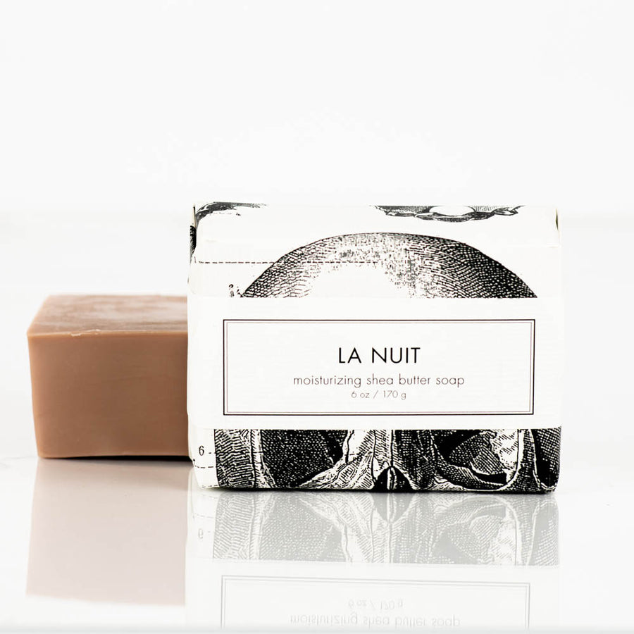 An unwrapped bar of brown natural soap sits next to a wrapped La Nuit Natural Shea Butter Bar Soap 6oz from Formulary55. The packaging of the La Nuit soap is white with black illustrations and text. The reflection of the soap items can be seen on the white surface.