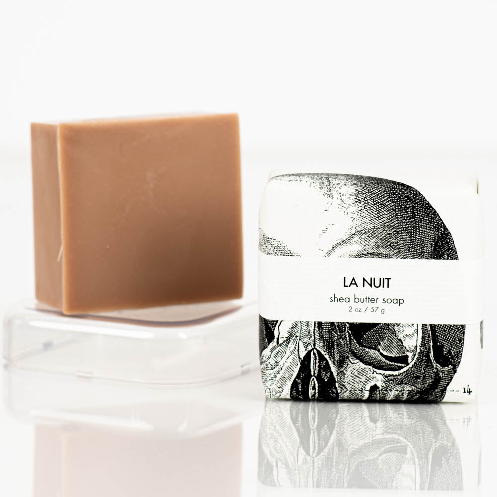 A brown natural shea butter bar soap is displayed on a clear soap dish next to its white and black packaging. The packaging features the text "Formulary55 la nuit petite shea butter natural bar soap 2 oz / 57 g" and an image of a skull. The background is white.