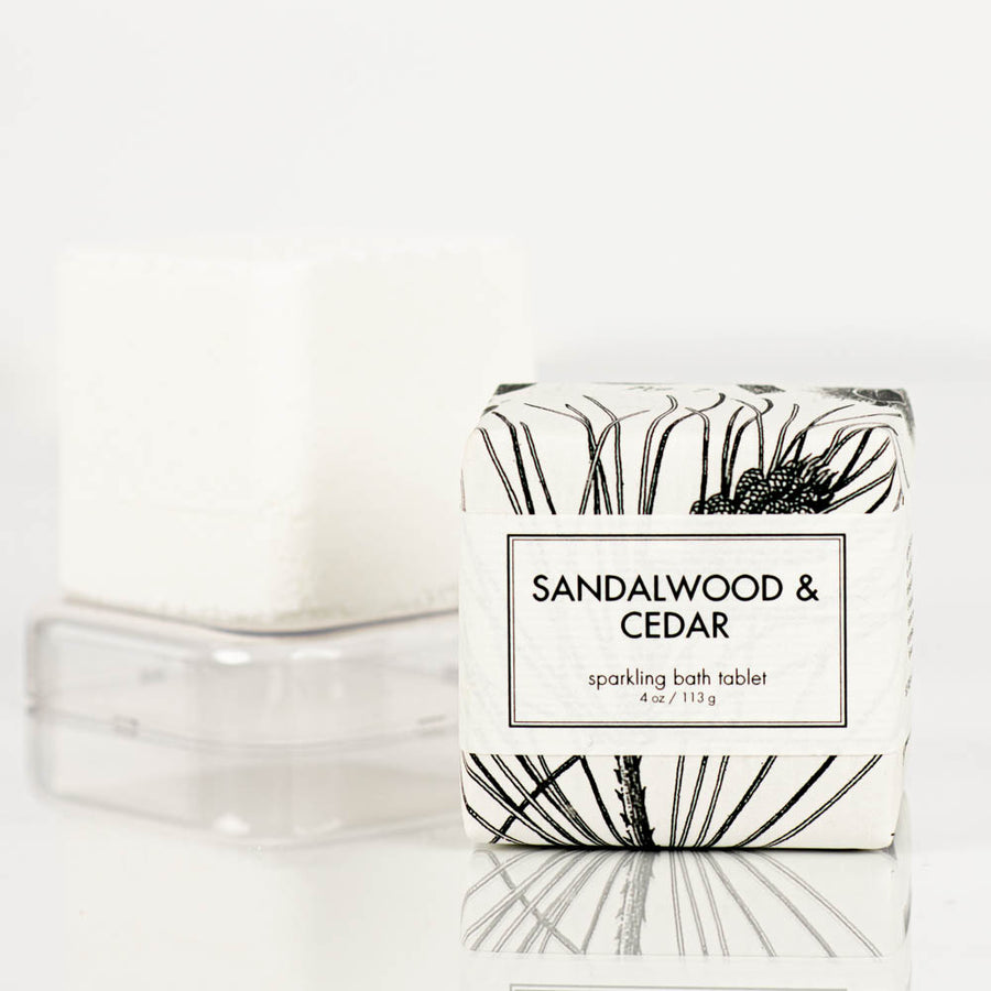 A Formulary55 "Sandalwood and Cedar" Sparkling Bath Bomb Tablet, weighing 4 oz (113 g), is elegantly wrapped in paper adorned with black botanical illustrations. It is displayed in front of an unwrapped, square-shaped white bath tablet on a clear stand.