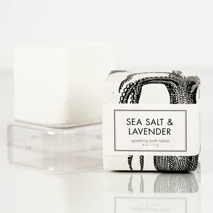 A Formulary55 sea salt and lavender Sparkling Bath Bomb Tablet wrapped in sleek white and black packaging sits prominently in the foreground. Behind it, an identical, unwrapped bath bomb tablet rests on a clear square tray, all set against a simple white backdrop.