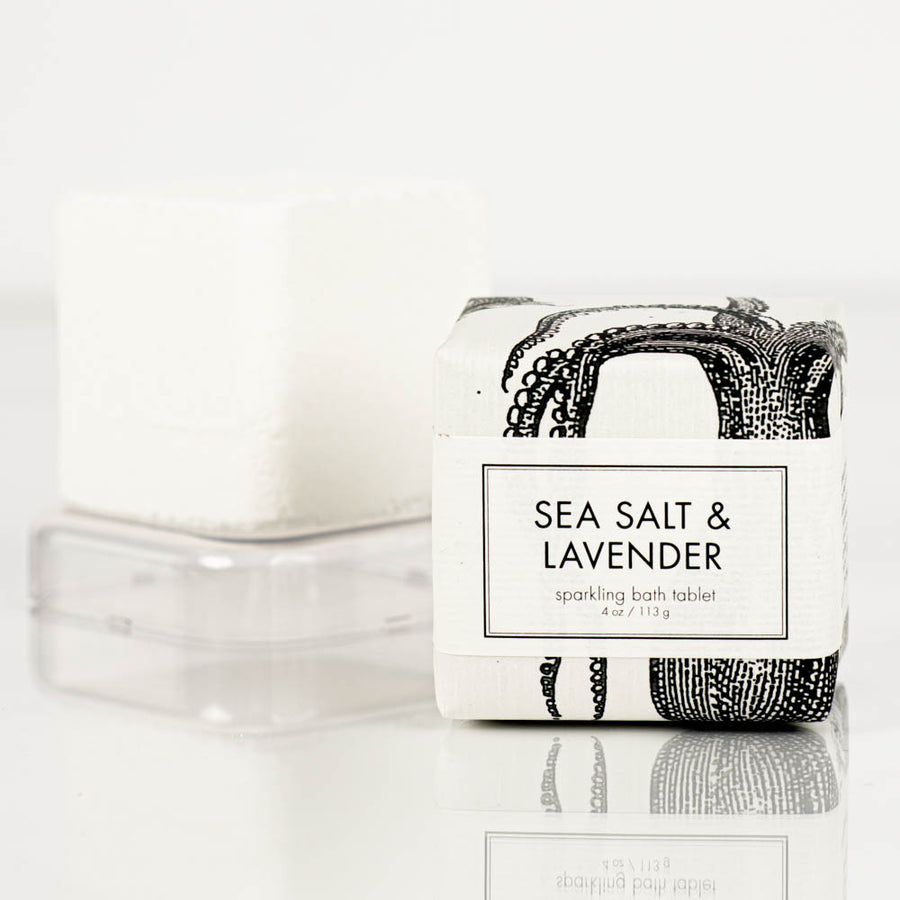 A Formulary55 sea salt and lavender Sparkling Bath Bomb Tablet wrapped in sleek white and black packaging sits prominently in the foreground. Behind it, an identical, unwrapped bath bomb tablet rests on a clear square tray, all set against a simple white backdrop.