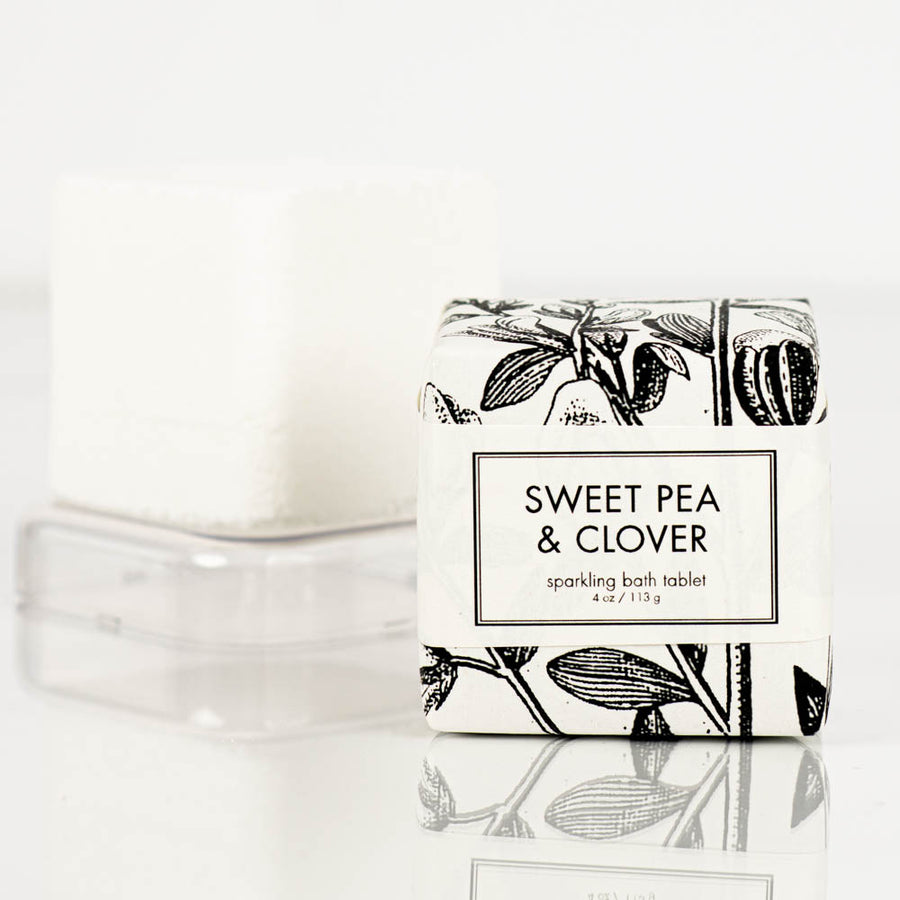 A white sparkling bath bomb tablet from Formulary55, labeled "sweet pea and clover" in black text, is shown wrapped in floral-patterned paper. An unwrapped tablet sits nearby on a clear plastic tray.