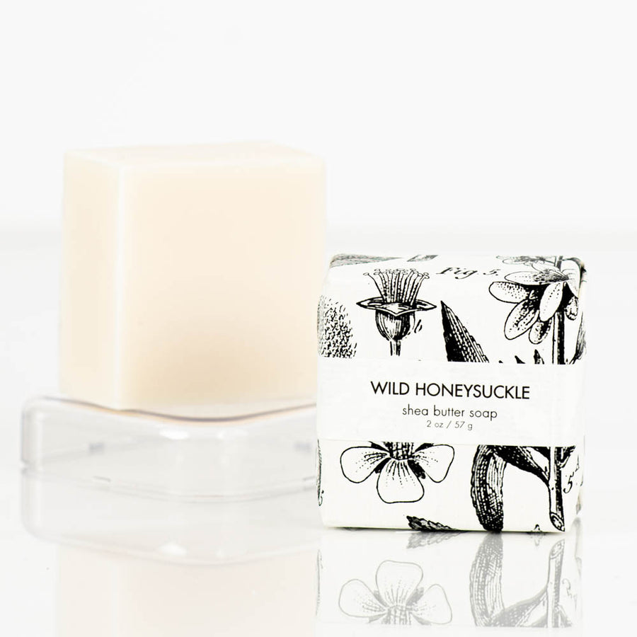 A beige bar of natural shea butter soap and its packaging are displayed on a white background. The soap sits on a clear soap dish while the packaging, labeled "Wild Honeysuckle Petite Shea Butter Natural Bar Soap" by Formulary55, features black floral illustrations.