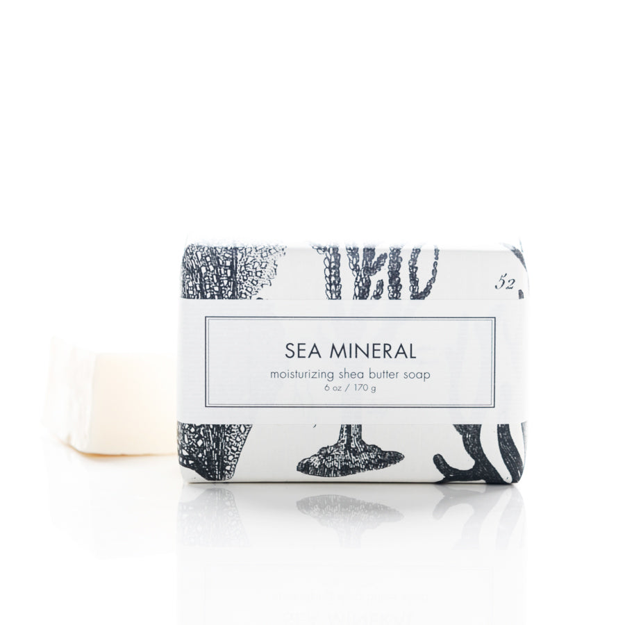 A white bar of "Sea Mineral" moisturizing shea butter soap is displayed against a white background. The soap is partially wrapped in white paper with black sea-themed illustrations. The wrapper has a label stating the soap's name and weight of 6 oz / 170 g.