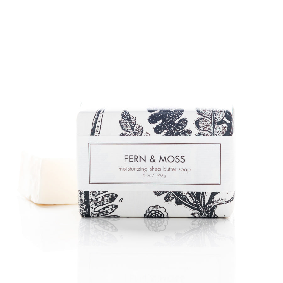 A rectangular bar of "Fern & Moss" moisturizing shea butter soap in minimalist packaging with black botanical illustrations. The 6 oz (170 g) soap bar is partially unwrapped, and a portion of the white soap is visible in the background on a white surface.