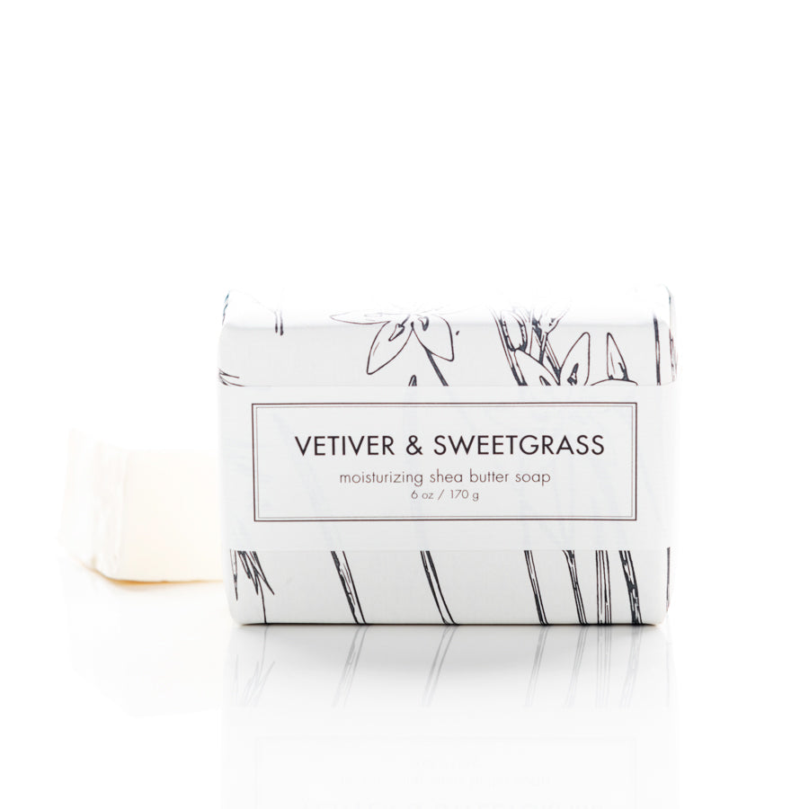 A bar of Vetiver & Sweetgrass moisturizing shea butter soap, wrapped in white paper with minimalist black botanical illustrations. The soap, partially unwrapped, is positioned on a clean white background. The label indicates a weight of 6 oz (170 g).