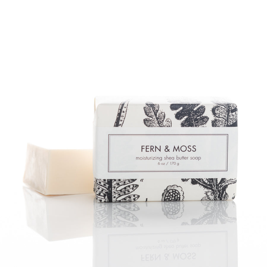 A rectangular bar of "Fern & Moss" moisturizing shea butter soap stands next to its partially visible cardboard packaging. The packaging is white with a black botanical illustration and text highlighting the soap's name and weight of 6 oz (170 g).