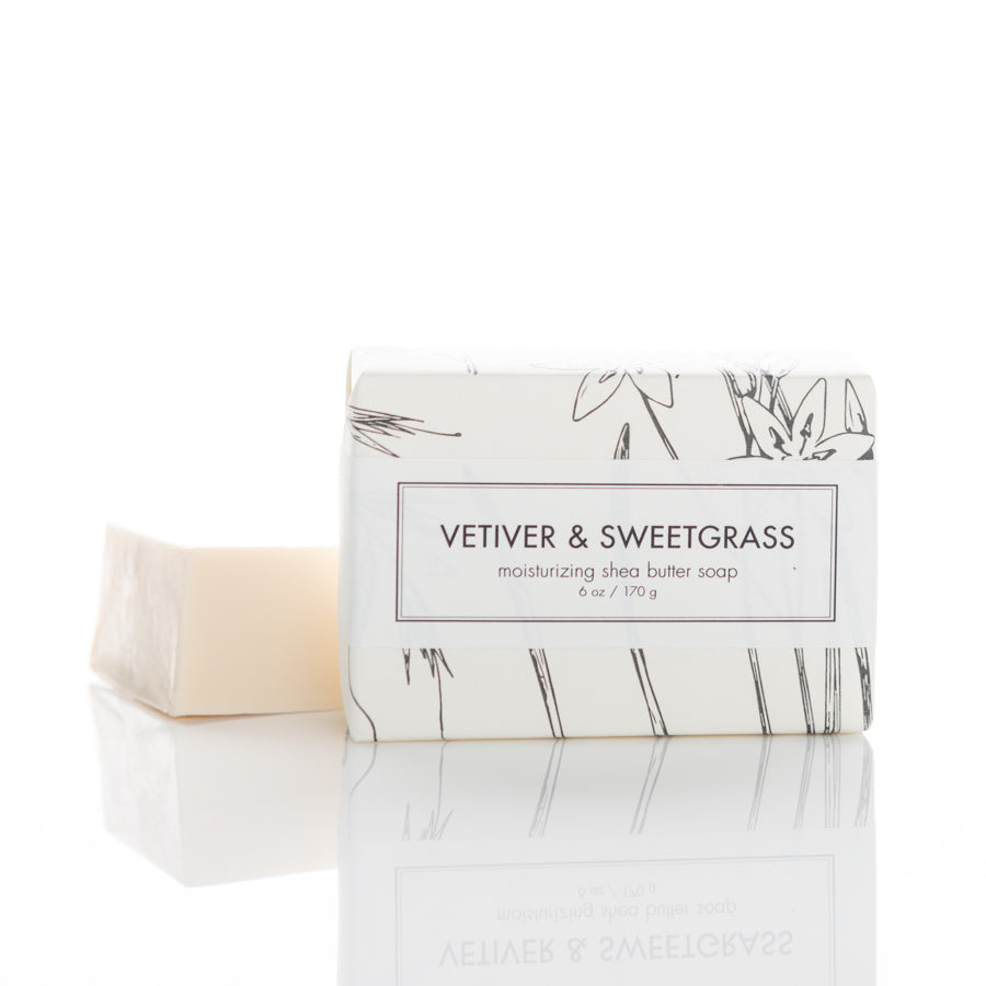 A bar of moisturizing shea butter soap labeled "Vetiver & Sweetgrass" is displayed. The soap, partly wrapped in white and gray botanical-themed paper, is positioned to the right of the unwrapped beige soap bar. The reflection is visible on the glossy surface.