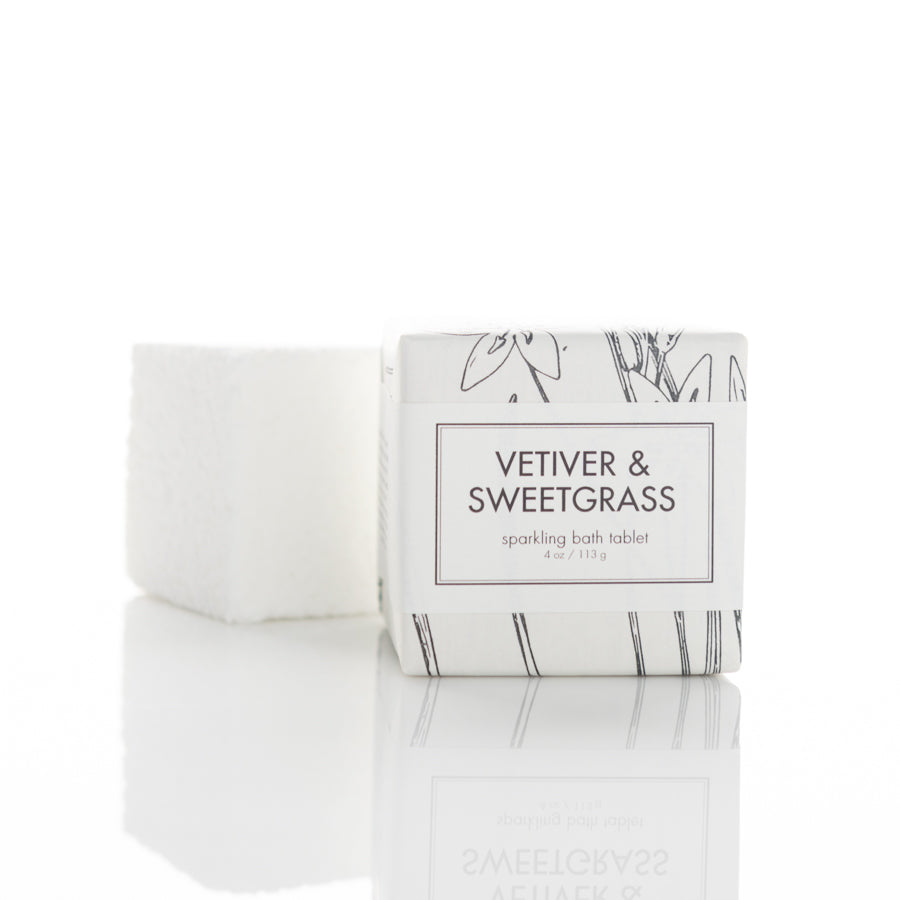 A white box labeled "Vetiver & Sweetgrass sparkling bath tablet" is placed beside a white, textured bath tablet. The box design features minimalistic black line art of florals. Both items are reflected on a glossy white surface.