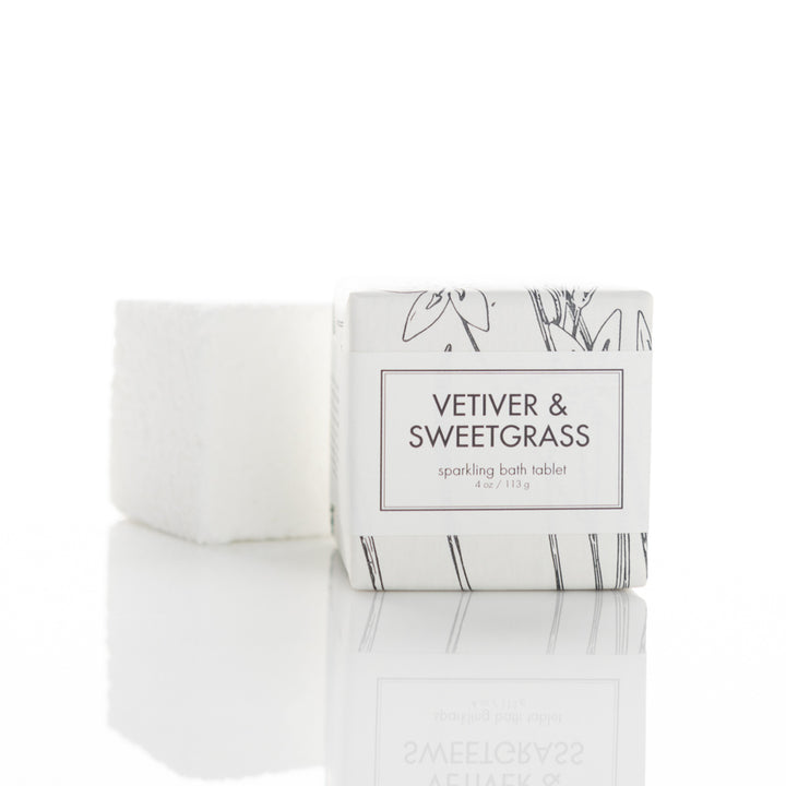 A white box labeled "Vetiver & Sweetgrass sparkling bath tablet" is placed beside a white, textured bath tablet. The box design features minimalistic black line art of florals. Both items are reflected on a glossy white surface.