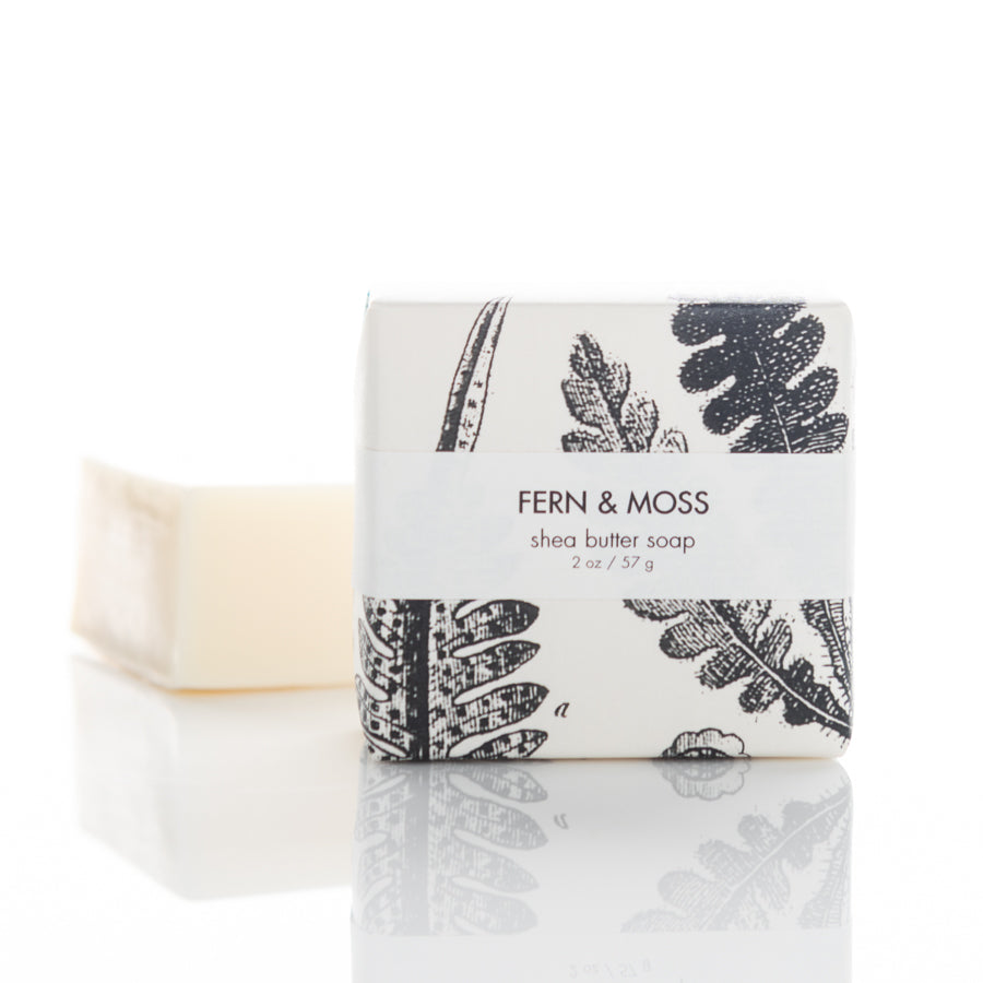 A bar of shea butter soap packaged in a white wrapper with botanical fern and moss illustrations. The wrapper has text that reads "FERN & MOSS shea butter soap 2 oz / 57 g." The unwrapped soap bar is placed adjacent to the wrapped one.