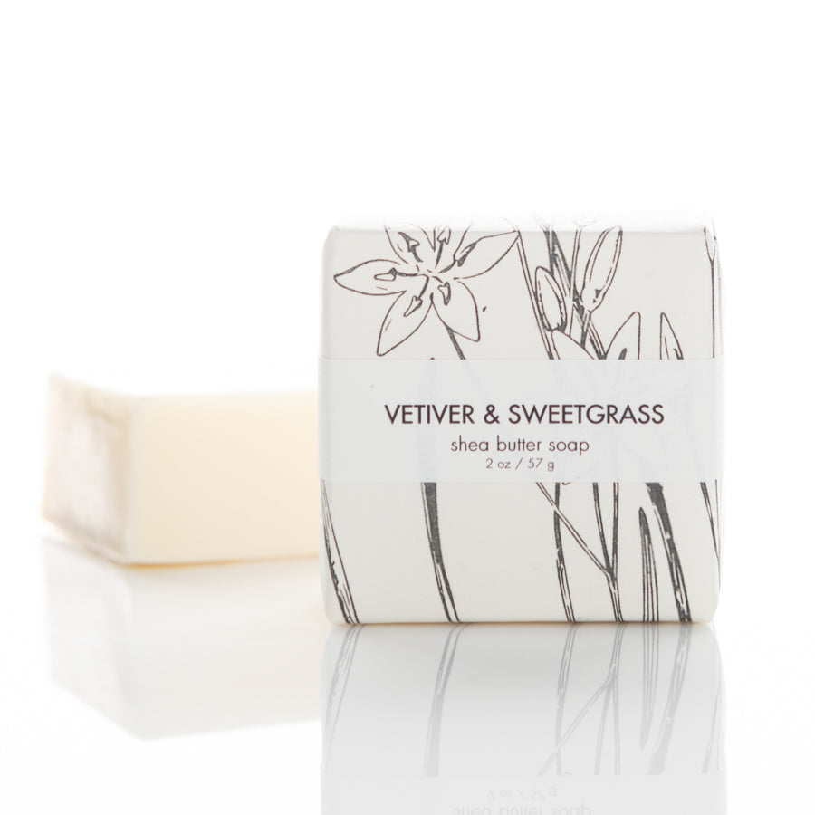 A bar of shea butter soap with vetiver and sweetgrass fragrance is wrapped in white paper adorned with minimalist plant drawings. The text "VETIVER & SWEETGRASS" and "shea butter soap 2 oz / 57 g" is printed on the front. The unwrapped soap is positioned alongside.