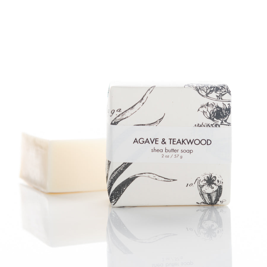 A bar of Agave & Teakwood shea butter soap in minimalist packaging with botanical illustrations. The soap is partially unwrapped, revealing the plain, beige bar inside. The text on the packaging indicates the soap weighs 2 oz (57 g).