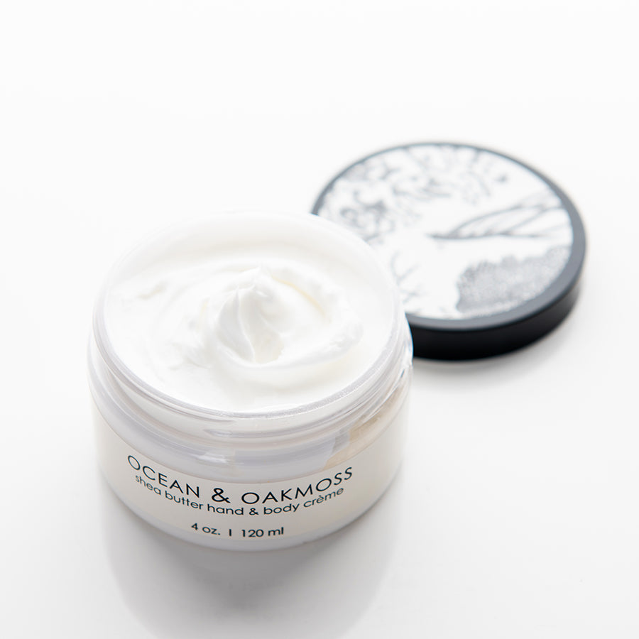 A white, round jar of shea butter hand and body creme labeled "Ocean & Oakmoss" with the lid off, showing the creamy product inside. The lid, partially visible and placed beside the jar, features a black and white illustration. The jar is 4 ounces (120 ml).