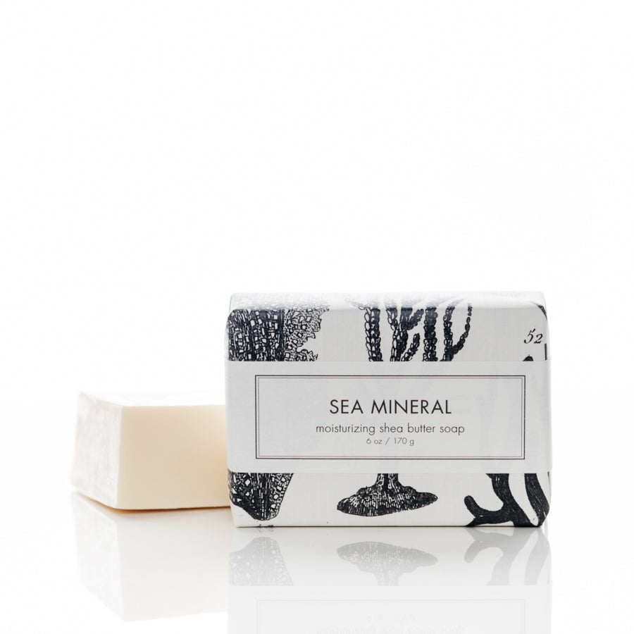 A bar of soap partially wrapped in white paper with black illustrations of sea elements. The label on the packaging reads "Sea Mineral moisturizing shea butter soap, 6 oz / 170 g." The soap itself is off-white.