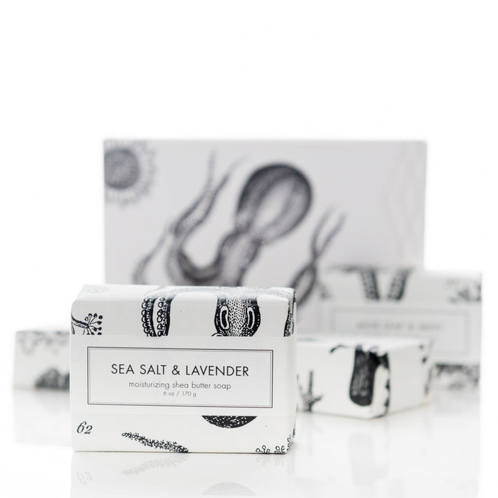 A small bar of soap labeled "Sea Salt & Lavender" is displayed in the foreground. The packaging has an elegant, monochrome design featuring sea creatures. Larger packages with a similar design are slightly blurred in the background.