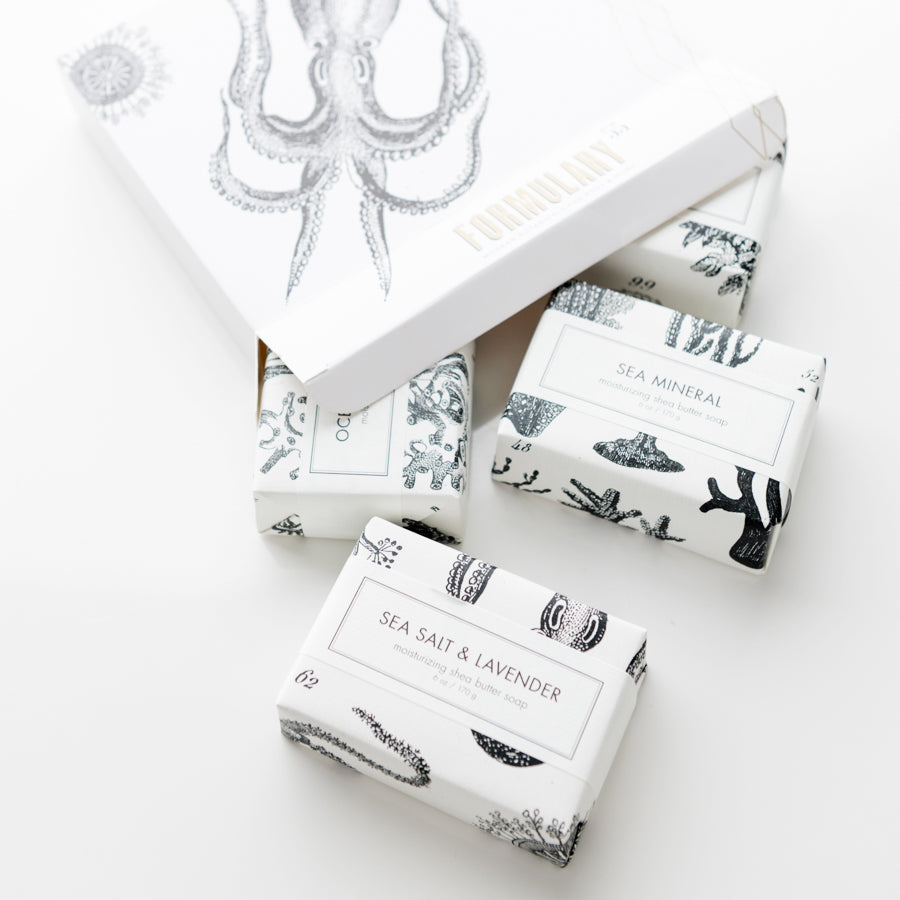 An arrangement of four sophisticatedly packaged soap bars lies on a white surface. The largest box features an illustrated octopus and reads "Finn & Co. FORMULARY 55." The smaller soap bars are labeled "WOOD," "SEA MINERAL," and "SEA SALT & LAVENDER.