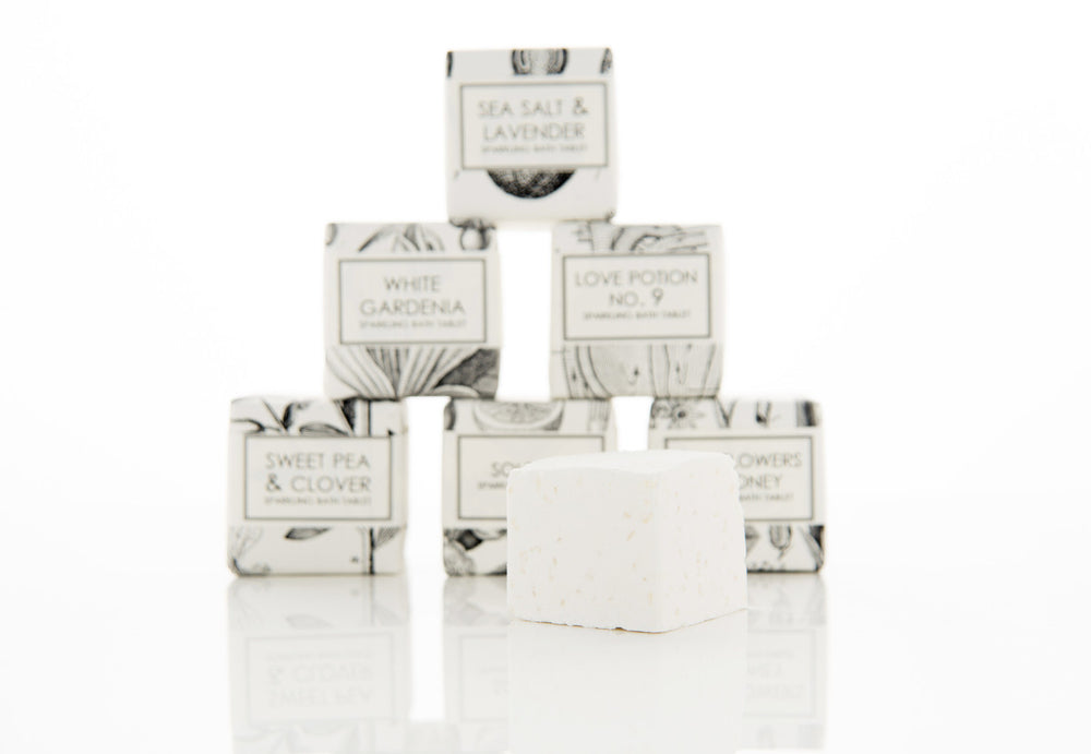 A pyramid arrangement of small, white, labeled soap boxes with a single unboxed soap bar in front. Soap scents include "Sea Salt & Lavender," "White Gardena," "Love Potion No. 9," "Sweet Pea & Clover," "Sugar Flower," and "Sunflower's Honey.