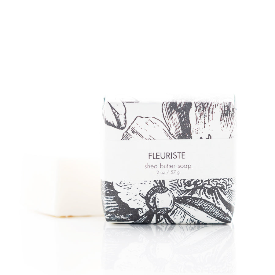 A bar of shea butter soap stands wrapped in elegant, floral-patterned paper labeled "Fleuriste." The soap, weighing 2 oz (57 g), is placed next to its wrapping, showing its plain white appearance. The background is white and minimalistic.