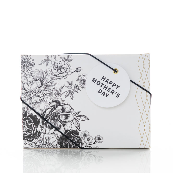 A rectangular gift box with a floral design in black and white is wrapped with black string. A circular white tag attached to the string reads "HAPPY MOTHER'S DAY" in black text. The box is placed on a reflective surface.