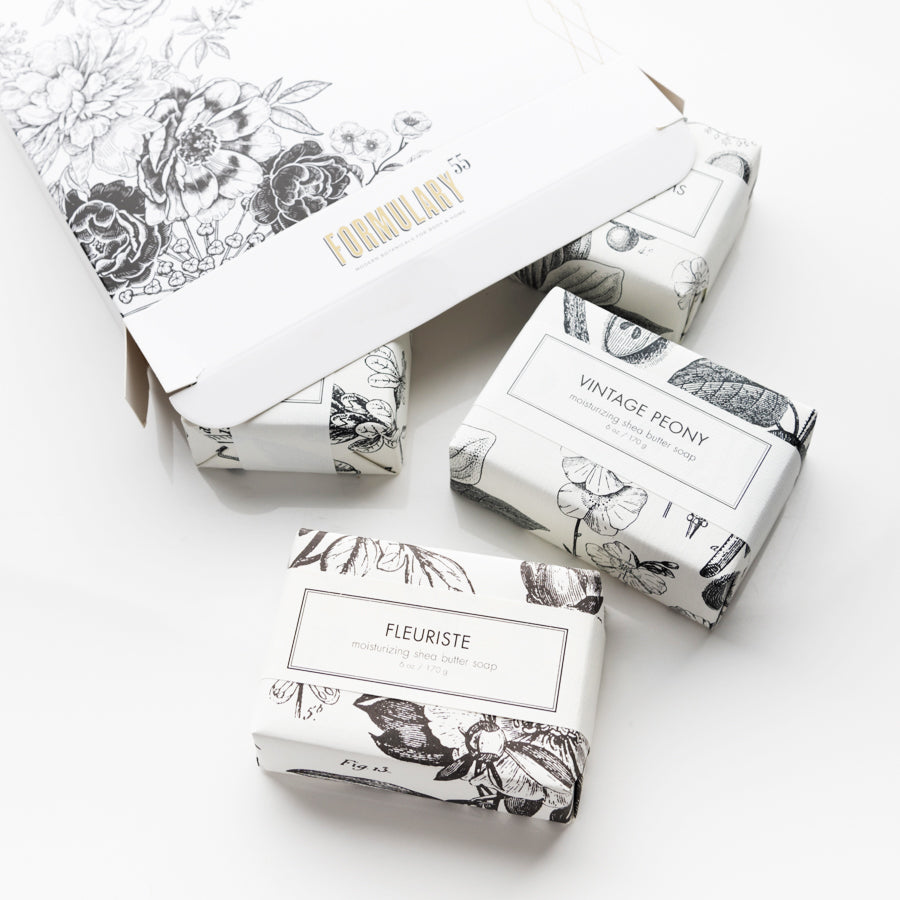 A white box with floral designs, labeled "Formulary 55," contains four elegant soap bars. The bars, in artistic floral packaging, are labeled "Vintage Peony," "Fleuriste," and others. The setup is minimalistic and clean, showcasing the products.