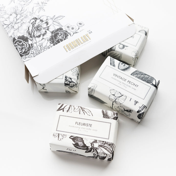 An open white box with a floral design reveals three individually wrapped soaps inside. The soaps are labeled "VINTAGE PEONY," "FLEURISTE," and another partially visible one with a floral print. The background is clean and minimalist.