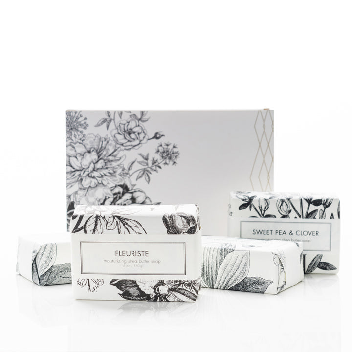 A set of three luxurious bar soaps with detailed botanical-themed packaging, placed in front of an elegantly designed box. One soap is labeled "Fleuriste", another "Sweet Pea & Clover", and the third is partially visible. The packaging features monochrome floral illustrations.