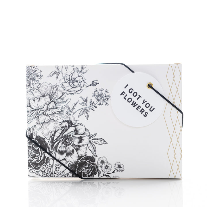 A rectangular gift box with an intricate black and white floral design. It is secured with a black string and has a circular white tag that reads, "I GOT YOU FLOWERS." The box reflects a minimalistic and elegant style.