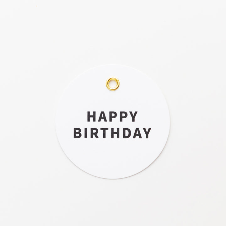 A round, white tag with a small gold grommet near the top center. The tag features the words "HAPPY BIRTHDAY" in bold, black capital letters. The simple design is set against a plain white background.