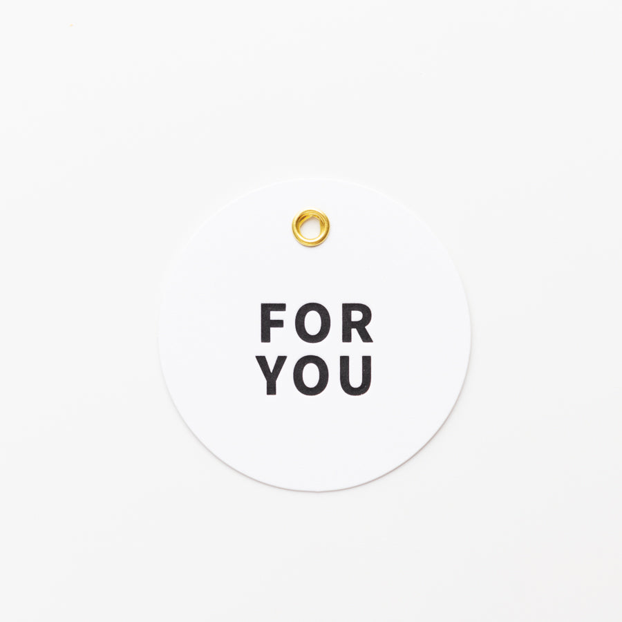 A white, circular tag with the words "FOR YOU" printed in bold black letters in the center. The tag has a single, small gold grommet near the top for attaching to a gift or item. The background is plain white.