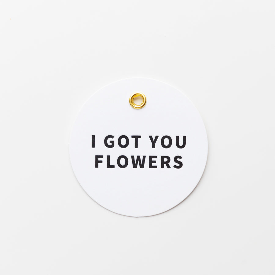 A round white tag with a small gold hole at the top center, featuring bold black text in the middle that reads "I GOT YOU FLOWERS.