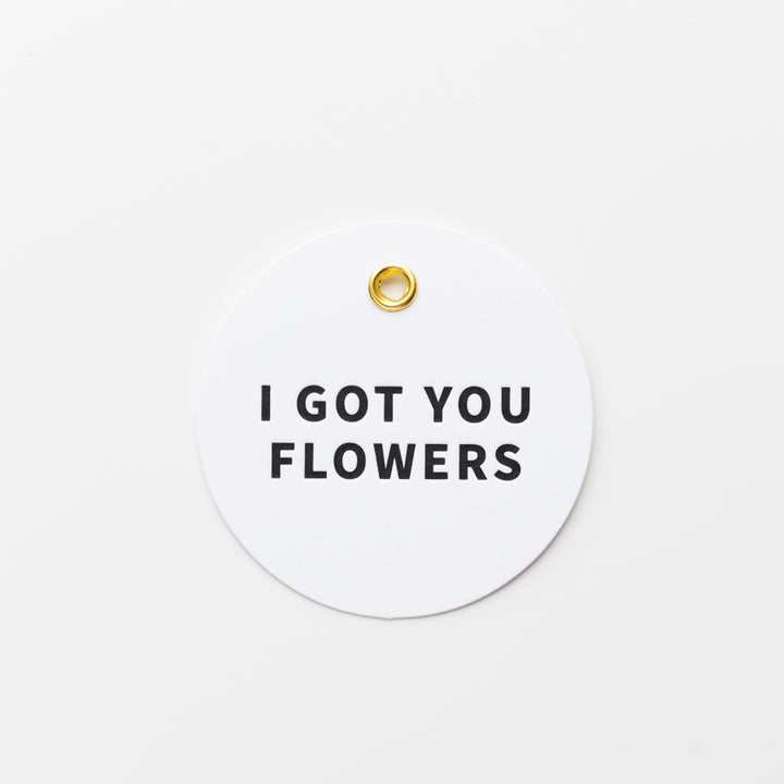 A round white tag with a small gold hole at the top center, featuring bold black text in the middle that reads "I GOT YOU FLOWERS.